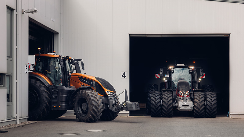 The image shows two large Valtra tractors, one orange and one black, positioned at the entrances of an agricultural dealership warehouse. The orange tractor is partially outside door number 5, while the black tractor is inside door number 4. The setting is clean and industrial, with visible numbers '4' and '5' marking the entrances. The overall mood is utilitarian and professional, with muted tones and strong lines.